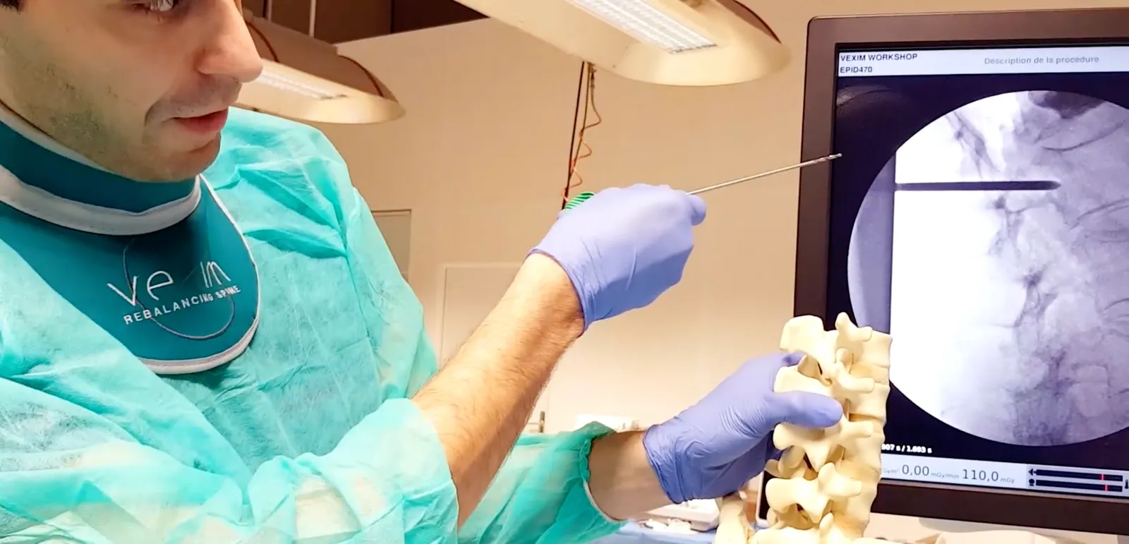 Un formateur en blouse chirurgicale montre, sur un modèle de colonne vertébrale, le trajet de l'instrument, devant l'écran de radiologie de la procédure